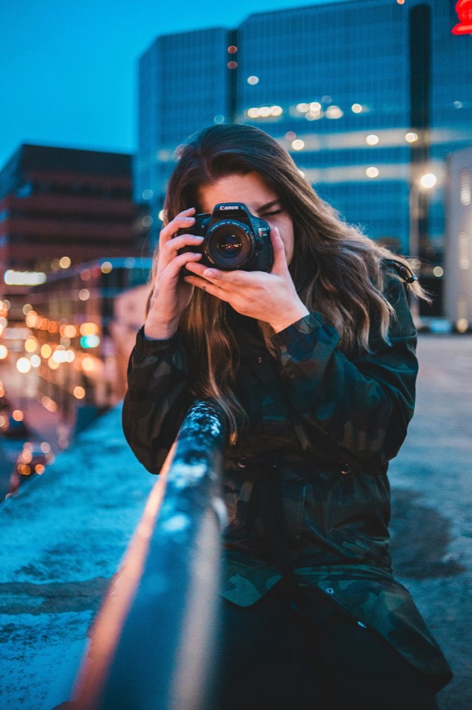 A photographer raising a camera to her eye on a city rooftop at dusk