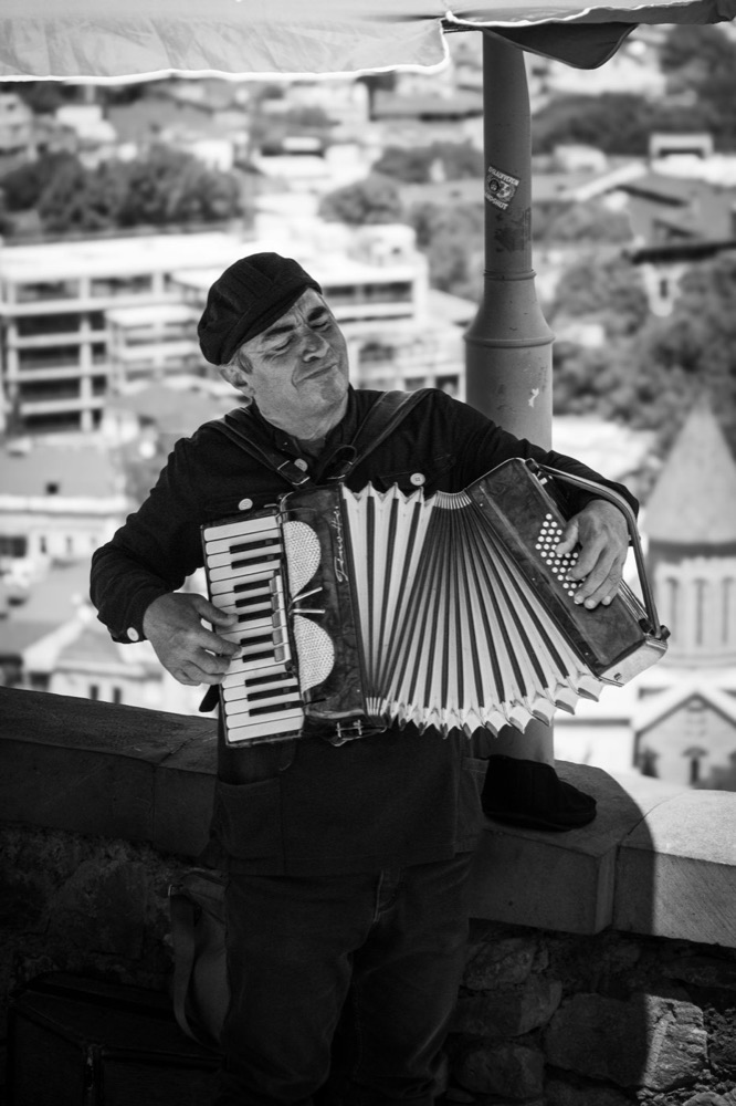 A man in a cap playing the accordion, eyes closed, above a hillside town